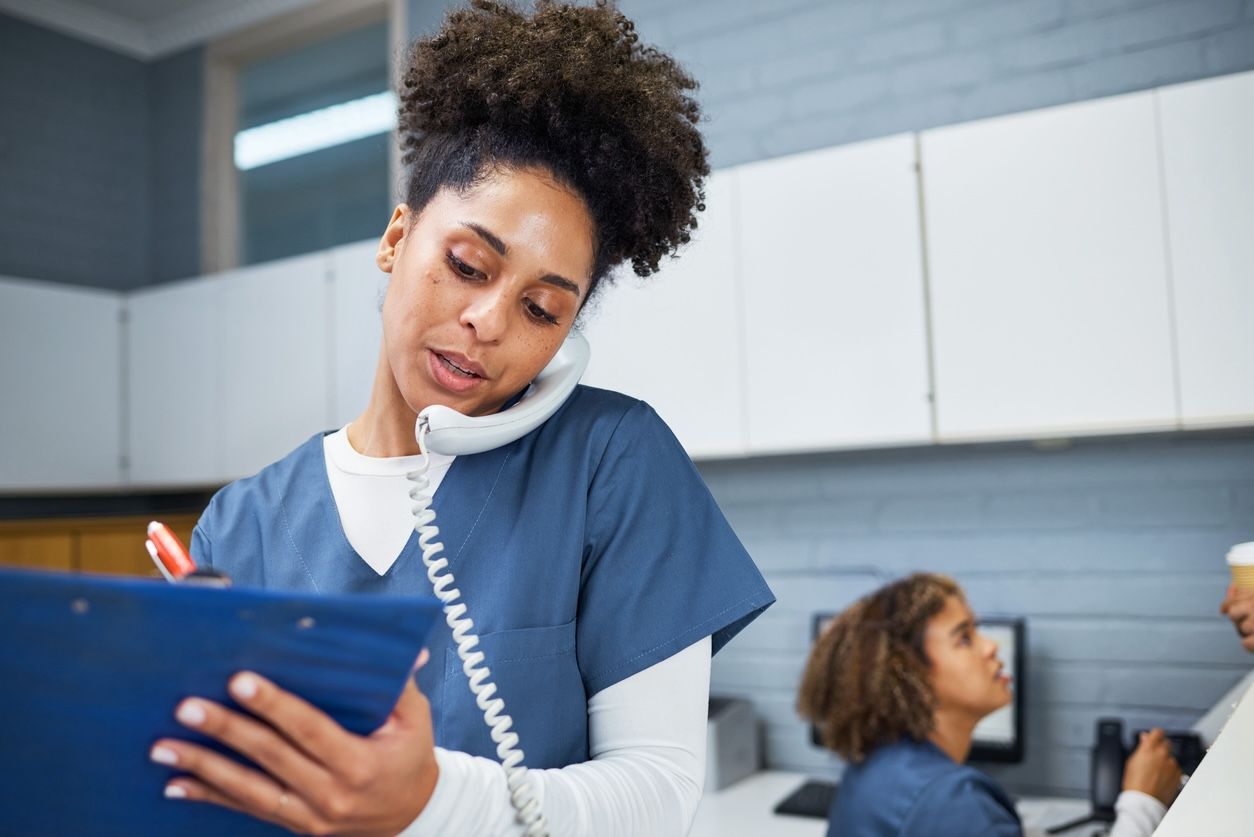 Female healthcare worker multitasking in a medical office—speaking on the phone while writing on a clipboard. The setting conveys a busy, professional environment, ideal for content related to time-saving services for medical professionals.